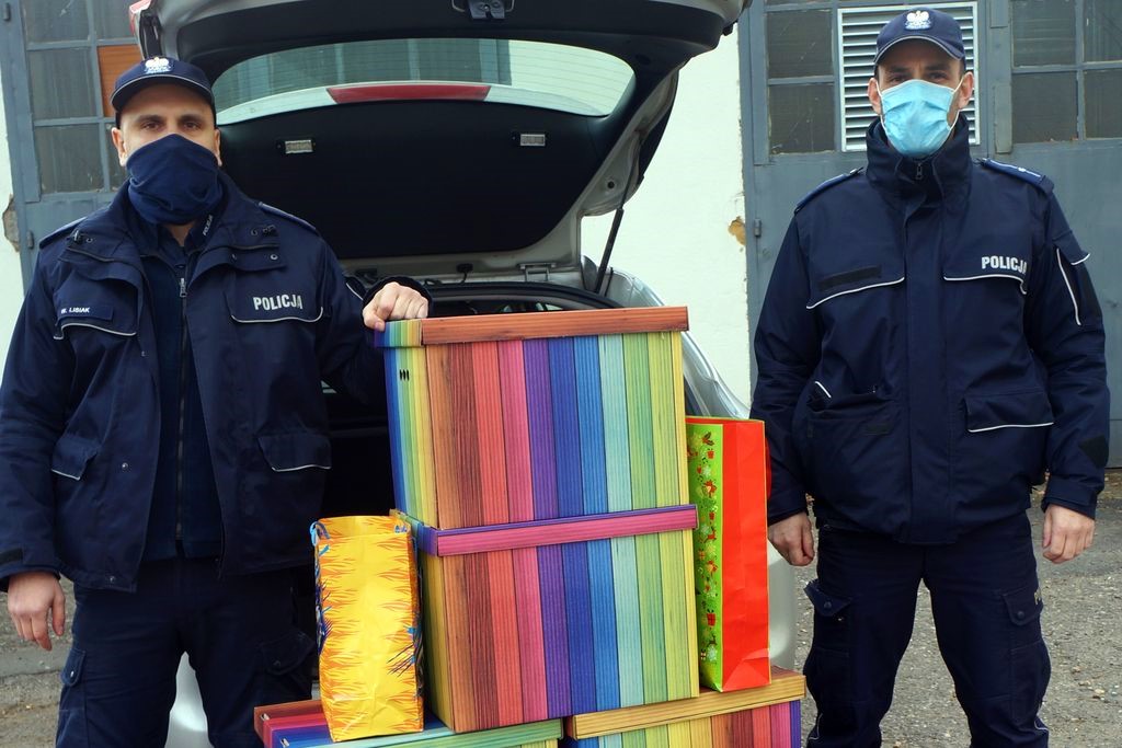 Policjanci stoją obok wielkich kolorowych kartonów z darami, a za nimi znajduje się otwarty bagażnik radiowozu.